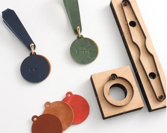 以下が含まれることがあります： 革細工のアイテムのコレクション。キーホルダーと木型が含まれています。キーホルダーは、革のストラップと金色の金具が付いた円形デザインです。革の色は、ネイビー、グリーン、ブラウン、レッドです。木型は革を切るためのものです。