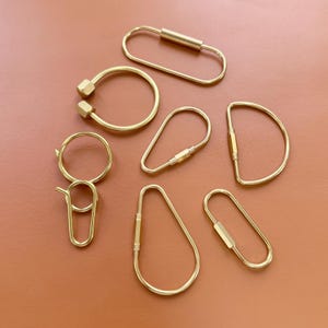 May include: A collection of brass keychains in various shapes, including oval, teardrop, and D-shaped designs. Some keychains feature screw-on closures, while others have a simple ring or clasp. The keychains are displayed on a brown surface.