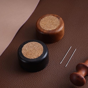 May include: Two wooden needle holders, one stained black and the other natural wood, each with a cork top. Two silver needles are next to the holders. The items are on a brown leather surface.