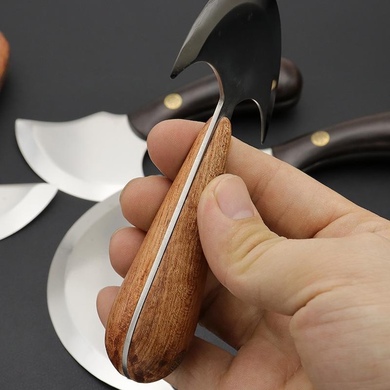 Op de afbeelding: Een mes met een gebogen lemmet en een houten handvat wordt in een hand gehouden. Het lemmet is zilverkleurig en het handvat is lichtbruin hout met een zilverkleurig accent. Andere messen zijn zichtbaar op de achtergrond.