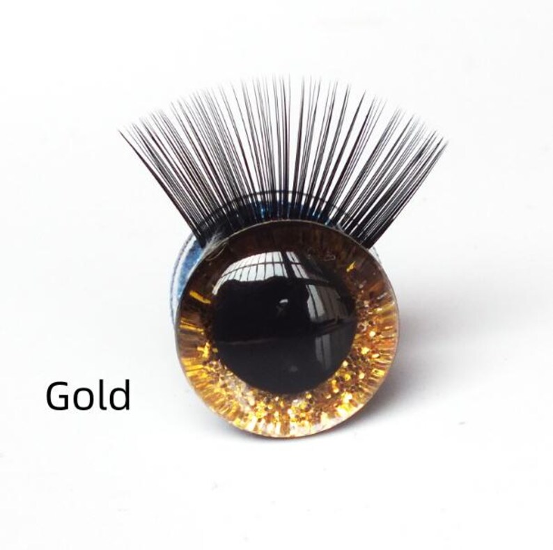 May include: A round, glass eye with a gold glitter rim and a black pupil. The eye has long, black eyelashes and the word "Gold" is printed on the left side. The eye is set against a white background.