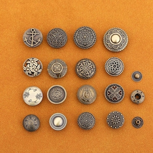 Könnte beinhalten: Eine Auswahl dekorativer Metallknöpfe in verschiedenen Größen und Designs. Die Knöpfe weisen aufwendige Muster auf, darunter florale, geometrische und nautische Motive. Die Farbpalette ist hauptsächlich Antikmessing und Bronze, mit einem orangefarbenen Hintergrund.