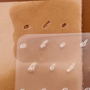 May include: Close-up of a leather surface with embossed letters 'a', 'b', and 'o'. A clear, curved plastic sheet overlays the leather, displaying the same letters in a white, cursive font. The leather is a light brown color.