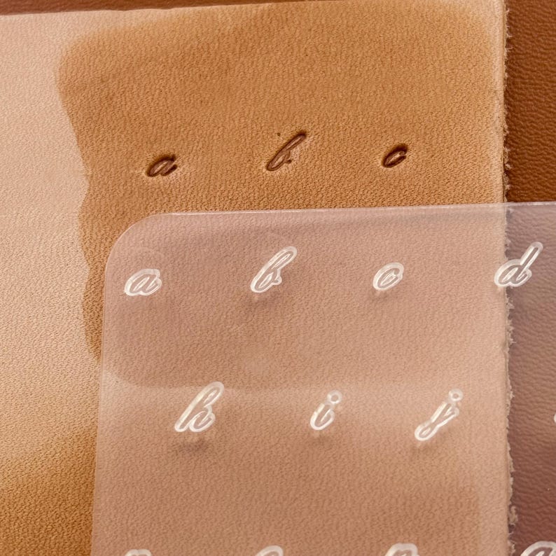 May include: Close-up of a leather surface with embossed letters 'a', 'b', and 'o'. A clear, curved plastic sheet overlays the leather, displaying the same letters in a white, cursive font. The leather is a light brown color.