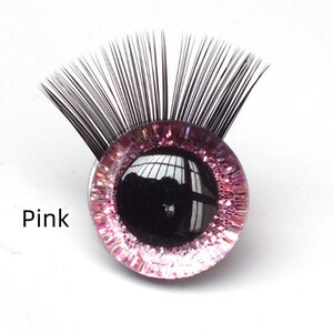 May include: A pink, round glass eye with black eyelashes. The eye has a black pupil and a pink glittery iris. The word "Pink" is printed in white on the left side of the eye.
