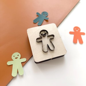 May include: A wooden die-cut tool with a metal outline of a gingerbread man, surrounded by three small, colorful gingerbread man cutouts in green, teal, and orange. The die is on a brown leather surface.