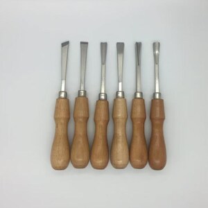 May include: A set of six wood carving chisels arranged in a row, each featuring a polished metal blade with a distinct tip shape and a smooth, light-coloured wooden handle. The tools are aligned vertically, showcasing varied cutting edges.