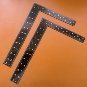 Puede incluir: Tres escuadras de carpintero de metal negro con marcas blancas, dispuestas sobre una superficie naranja. Las escuadras tienen medidas en centímetros. Estas herramientas están diseñadas para mediciones precisas en ángulo recto.