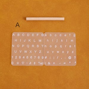 May include: A clear, rectangular alphabet and number stamp set with uppercase and lowercase letters, numbers, and symbols. A small wooden dowel is above the stamp set. The background is a solid orange color.