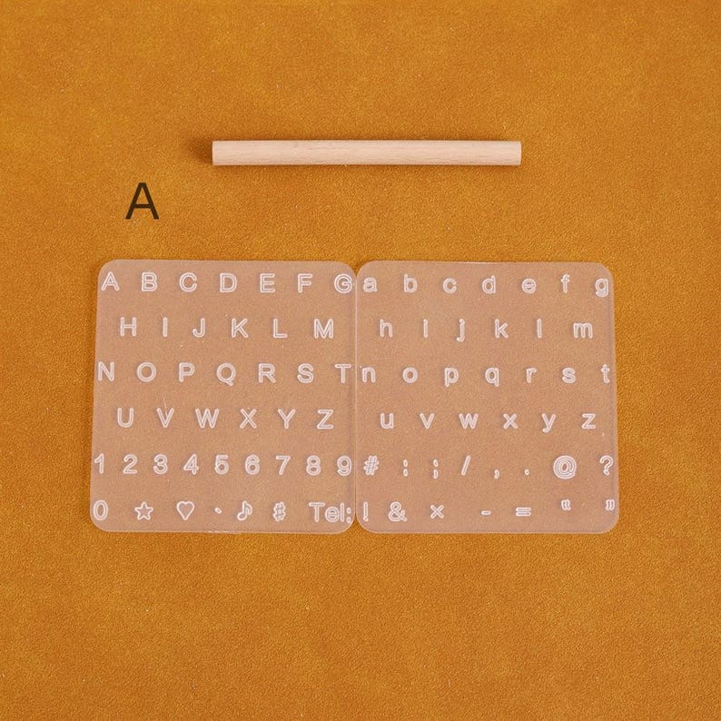 May include: A clear, rectangular alphabet and number stamp set with uppercase and lowercase letters, numbers, and symbols. A small wooden dowel is above the stamp set. The background is a solid orange color.