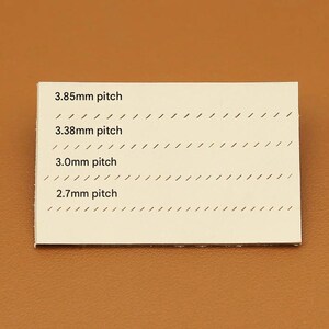 May include: A rectangular, off-white card with four lines of text indicating stitch pitch measurements: 3.85mm, 3.38mm, 3.0mm, and 2.7mm. Each line is accompanied by a series of angled, black lines. The card is set against a brown background.
