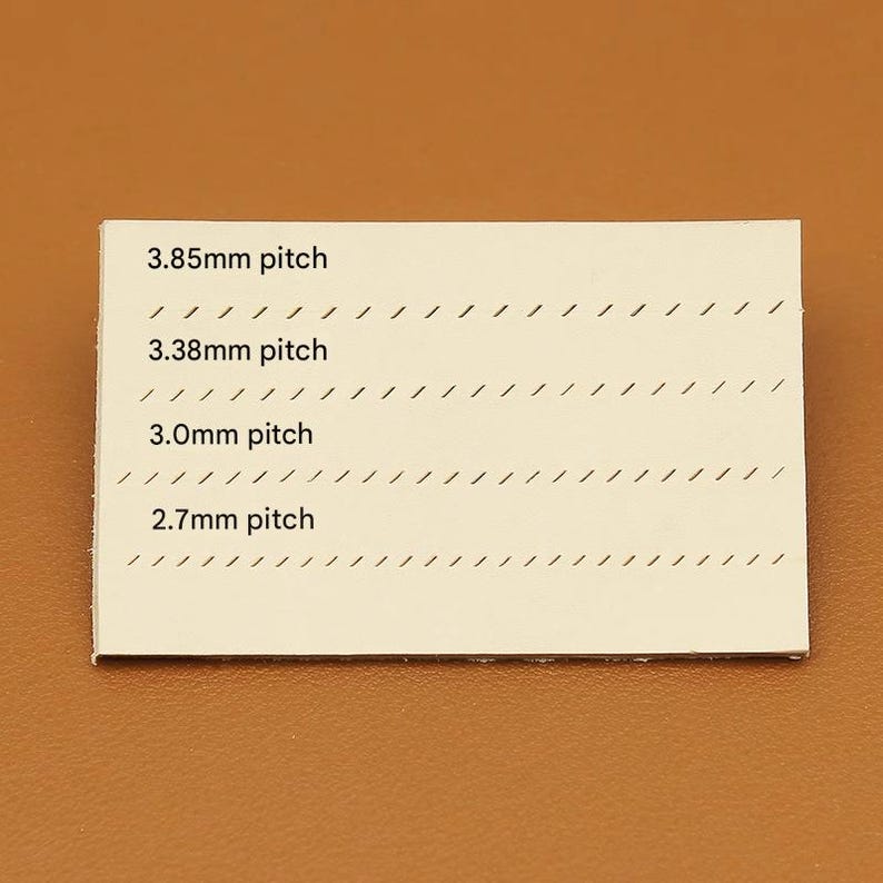 May include: A rectangular, off-white card with four lines of text indicating stitch pitch measurements: 3.85mm, 3.38mm, 3.0mm, and 2.7mm. Each line is accompanied by a series of angled, black lines. The card is set against a brown background.