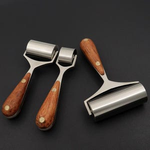 Op de afbeelding: Drie handrollen van metaal en hout. De rollen hebben een roestvrijstalen cilinder en een houten handvat met een messing accent. De handvatten zijn warmbruin en het metaal is zilverkleurig. Geplaatst op een zwarte ondergrond.