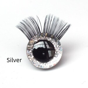 May include: A decorative eye-shaped embellishment with a black pupil and silver glitter detailing. The eye is surrounded by a clear, circular frame and topped with black, feathery lashes. The word "Silver" is printed below the eye.