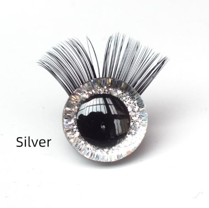 May include: A decorative eye-shaped embellishment with a black pupil and silver glitter detailing. The eye is surrounded by a clear, circular frame and topped with black, feathery lashes. The word "Silver" is printed below the eye.