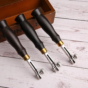 May include: Three leather edge creasers with dark wooden handles, brass accents, and adjustable metal blades. The tools are designed for leathercraft and feature a screw for adjusting the blade width. The handles are a dark brown and black.