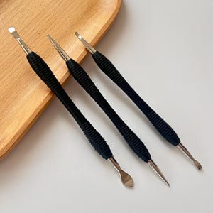 Puede incluir: Tres herramientas para uñas con mangos negros texturizados y puntas de metal plateado. Las herramientas tienen diferentes formas de punta, incluyendo una cuchara, un extremo puntiagudo y un extremo plano y en ángulo. Las herramientas están dispuestas sobre una bandeja de madera.