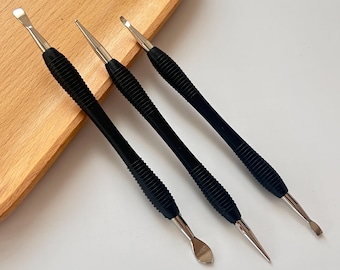 May include: Three nail tools with black, textured handles and silver metal tips. The tools have different tip shapes, including a scoop, a pointed end, and a flat, angled end. The tools are arranged on a wooden tray.