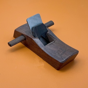 Puede incluir: Un cepillo de carpintero de madera marrón oscuro con una hoja metálica y una cuña de madera oscura asegurada en el cuerpo. La herramienta incluye un mango horizontal de madera que se extiende por los lados, con un acabado suave y pulido.