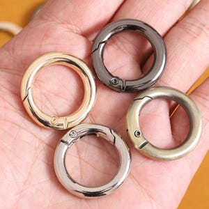 Pode incluir: Quatro anéis redondos de metal com mola nas cores dourado, cinza chumbo, prata e latão antigo. Cada anel tem uma abertura articulada com um pequeno fecho de parafuso. Os anéis são mostrados em uma mão.