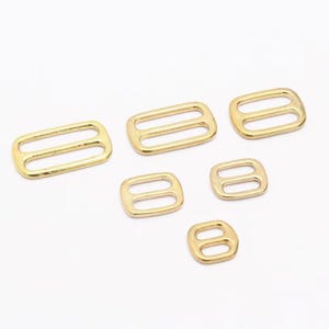 Op de afbeelding: Een verzameling goudkleurige metalen schuifregelaars in verschillende maten. Elke schuifregelaar heeft een rechthoekige of vierkante vorm met twee parallelle sleuven. De schuifregelaars zijn gerangschikt op een wit oppervlak.