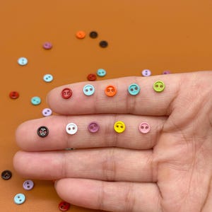 May include: A collection of small, colorful buttons in various shades, including red, blue, orange, and green. The buttons are round with two holes and a smiley face design. Some buttons are held on a person's fingers.