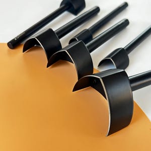 May include: A collection of black leathercraft tools with curved blades and cylindrical handles. One tool is marked with "25mm". The tools are arranged on a white and orange surface.