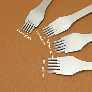 May include: Four silver leather stitching tools with different pitch sizes: 2.7mm, 3.0mm, 3.38mm, and 3.85mm. The tools are arranged on a brown surface, showcasing their various prong configurations for leathercraft.