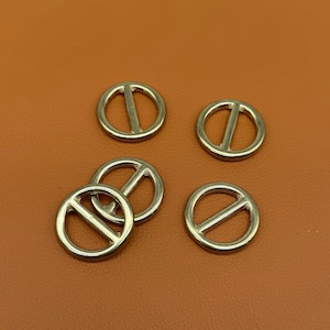 Può includere: Cinque anelli per sciarpa in metallo color oro, ciascuno di forma circolare con una barra diagonale al centro. Gli anelli sono disposti su una superficie marrone, creando una presentazione semplice ma elegante. Accessori per sciarpe.