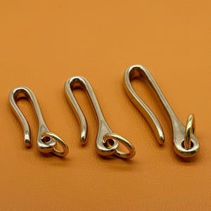 May include: Three metal keychains with a hook design. The keychains are silver-colored with a gold-colored ring at the bottom. The keychains are arranged from smallest to largest, with a curved hook shape.
