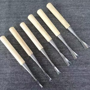 May include: Six wood carving tools with light-colored wooden handles and silver-colored metal blades. The blades have a curved shape. The tools are arranged diagonally on a gray surface.