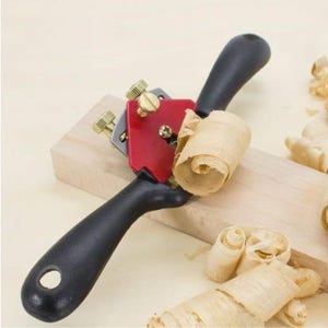 Könnte beinhalten: Ein Schweifhobel für die Holzbearbeitung mit zwei schwarzen ergonomischen Griffen, einem roten Metallkörper in der Mitte und goldenen Einstellschrauben. Das Werkzeug liegt auf einem hellen Holzblock mit mehreren lockigen Holzspänen daneben.