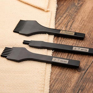 May include: Three black leathercraft tools with the text "2.7 mm" on each handle. The tools are arranged on a wooden surface with a beige fabric in the background. The tools have different shaped heads.