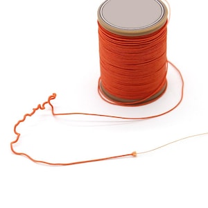 May include: A spool of bright orange thread with a silver top and a brown cardboard base. A length of the thread extends from the spool, with a small knot at the end. The thread is a vibrant, solid color.