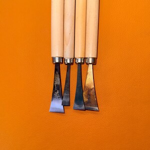 May include: Four wood carving tools with light-colored cylindrical wooden handles and metal ferrules. The blades are made of dark steel with varying widths and shapes, including flat and angled edges, showing signs of use and metallic patina.
