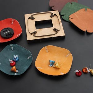 Puede incluir: Un surtido de suministros para manualidades en cuero y productos terminados. Incluye una matriz de madera, varias piezas de cuero en varios colores y pequeños platos de cuero con costuras decorativas. Se ve una tapa de objetivo Canon.