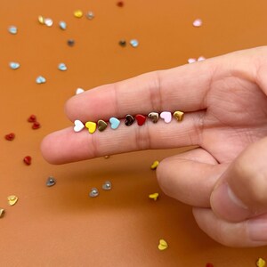 Peut inclure: Une collection de petites boucles d'oreilles en forme de cœur de différentes couleurs : blanc, jaune, bleu, marron, rouge et rose. Les clous sont disposés sur un doigt sur un fond marron, avec d'autres cœurs dispersés.