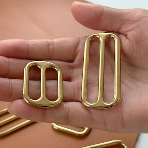 May include: Two gold-colored metal buckles with a rectangular shape and three slots. One buckle is smaller and more rounded. The buckles are likely for use in bags, belts, or other accessories.