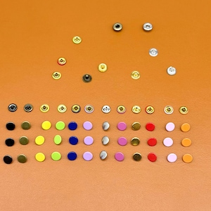 May include: A collection of various metal snap fasteners in multiple colors, including black, gold, silver, and a range of bright hues. The snaps are arranged on a solid orange background, showcasing their different finishes and styles.