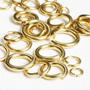 May include: A collection of gold-colored jump rings of various sizes, some open and some closed. The rings are scattered across a white surface, with a focus on the metallic sheen and circular shape. Ideal for jewelry making and crafting.
