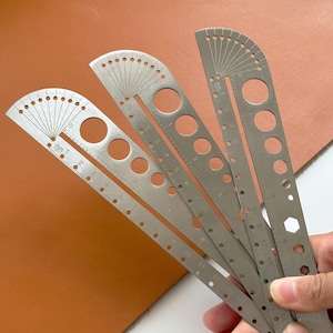 Peut inclure: Ensemble de règles en acier inoxydable de différentes formes. Chaque règle comporte des mesures en centimètres et en pouces, ainsi que des découpes circulaires et hexagonales. Les règles sont présentées sur un fond marron.