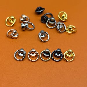 Puede incluir: Una colección de remaches con anillo de metal en varios acabados metálicos, incluyendo oro, plata, bronce y gris plomo. Cada pieza consta de un anillo circular unido a una pequeña base de perno redondeada para fijar en cuero o tela.