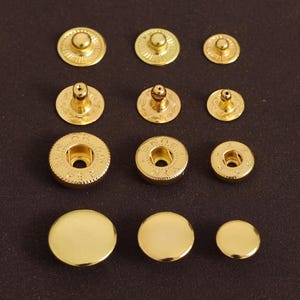 以下が含まれることがあります： さまざまなサイズの金色の金属製スナップファスナーのコレクション。スナップには、キャップ、ポスト、ソケットなどのさまざまなコンポーネントが含まれています。留め具は濃い茶色の表面に配置され、光沢のある金属仕上げが施されています。