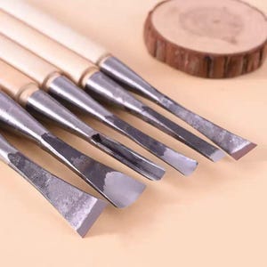 May include: A set of six wood carving chisels with wooden handles and metal blades. The blades have various shapes for different carving techniques. A small, round wooden slice is in the background.