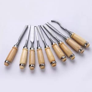 Könnte beinhalten: Eine Sammlung von Holzmeißeln mit Holzgriffen und Metallklingen. Die Meißel sind fächerförmig angeordnet und zeigen ihre verschiedenen Klingentypen. Die Griffe sind hellbraun und die Metallklingen sind silberfarben.