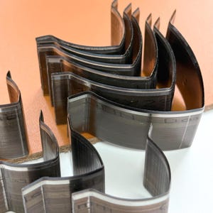 May include: A collection of steel leather cutting dies, arranged in a curved pattern. The dies have a dark, metallic finish with sharp edges, designed for precision leather crafting. The image shows the tools on a neutral background.