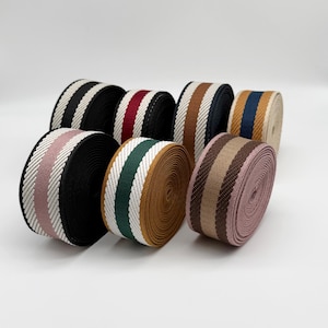 Puede incluir: Una colección de rollos de cinta tejida colorida. Los rollos muestran varias combinaciones de colores, incluyendo negro y blanco, rojo y negro, marrón y blanco, y verde y blanco. Las cintas están bien enrolladas y listas para proyectos de manualidades o diseño.