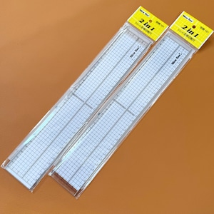 Op de afbeelding: Twee doorzichtige plastic linialen, elk 30 cm lang, met een rasterpatroon en meetmarkeringen. De linialen zijn verpakt in doorzichtige plastic hoezen met gele en witte labels met de tekst "2 in 1" en "30cm".
