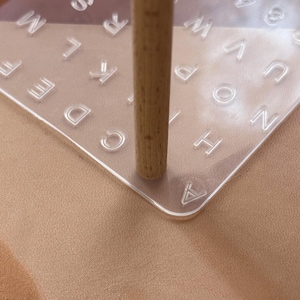 May include: A clear, square acrylic stamp with embossed letters and a wooden handle. The stamp is partially pressed onto a tan surface, with the letters visible. The wooden handle is cylindrical and light brown.