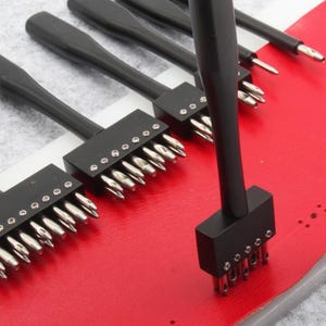May include: A set of black leather working tools with silver metal tips. The tools are arranged on a red leather surface, with a gray background. The tools appear to be used for punching holes in leather.